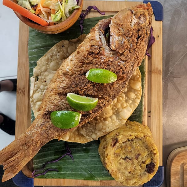 Fried whole fish with lime wedges, mofongo, salad, and crispy flatbread on a banana leaf.
